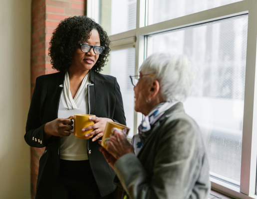 A younger and older adult holding mugs by a window.