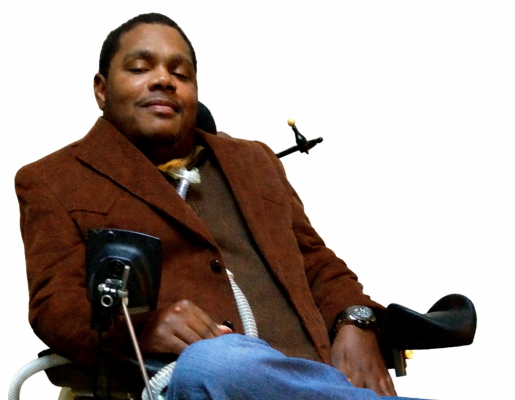 Photo of Travar Pettway sitting in a motorized wheelchair wearing a brown suit jacket and jeans.