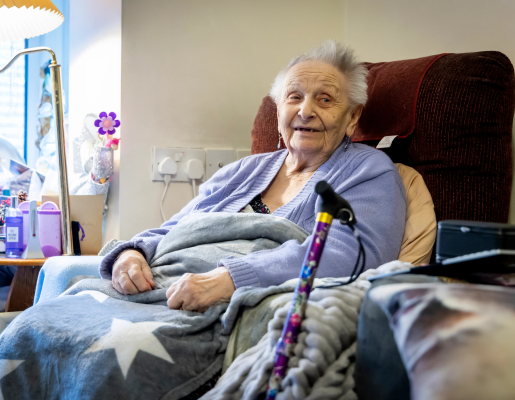 Older woman sitting on a couch with blankets. Her cane sits against the couch's armrest.