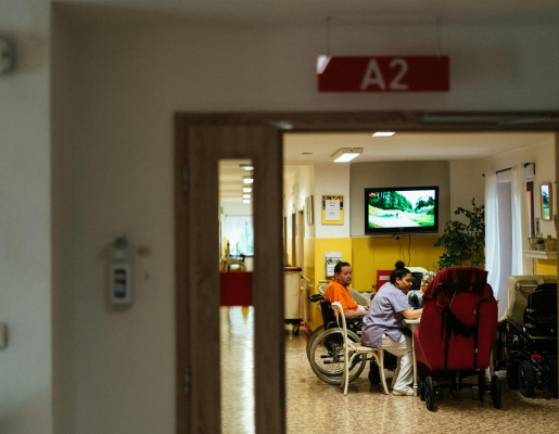 View through a doorway of a group of older adults at a table in a nursing home. One person is in a wheelchair and another wearing scrubs.