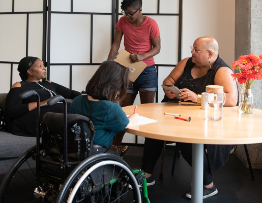 A group of black and brown adults sitting by a table. One of them is in a wheelchair. 