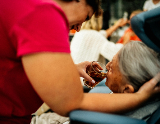 A caretaker standing over an older adult, giving them a drink from a glass while they lay in bed. 