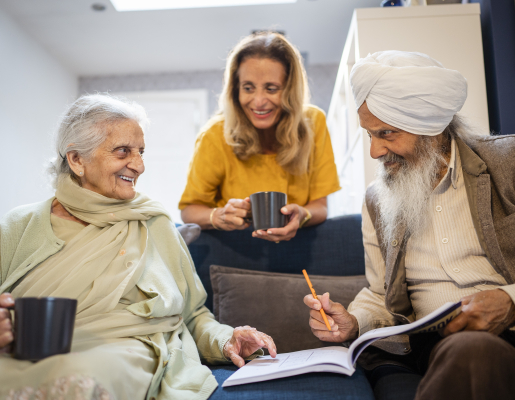 Two older adults sitting on a couch, with a younger adult behind them. They are smiling at each other while holding mugs or writing in a book.