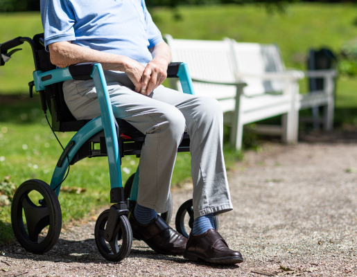 An older adult, sitting in a walker, outside by a bench.