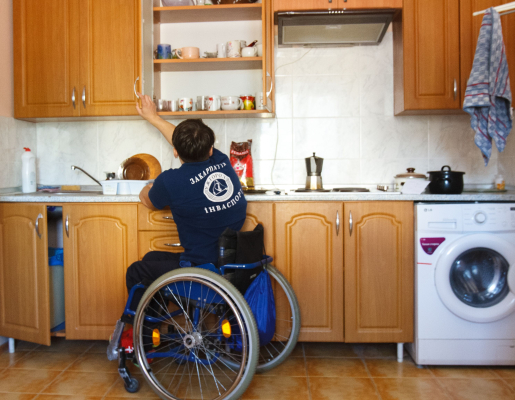 A person in a wheelchair reaching up to get something from a kitchen shelf. 
