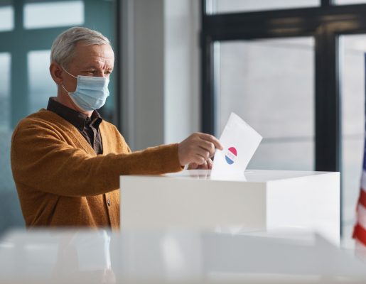 Image of an older man wearing a medical mask putting his ballot into a ballot box. 