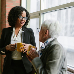 A younger and older adult holding mugs by a window.