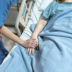 Image of a patient in a bed and a nurse holding their hand.