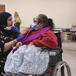 Younger woman wearing a hijab and kneeling, talking to an older woman in a wheelchair.