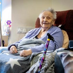 Older woman sitting on a couch with blankets. Her cane sits against the couch's armrest.