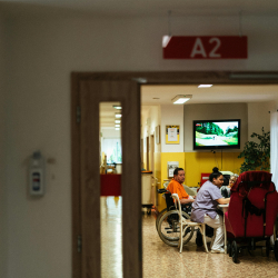 View through a doorway of a group of older adults at a table in a nursing home. One person is in a wheelchair and another wearing scrubs.