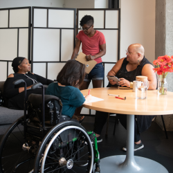 A group of black and brown adults sitting by a table. One of them is in a wheelchair. 