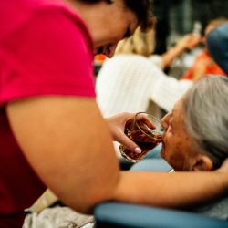 A caretaker standing over an older adult, giving them a drink from a glass while they lay in bed. 