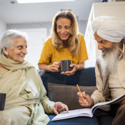 Two older adults sitting on a couch, with a younger adult behind them. They are smiling at each other while holding mugs or writing in a book.