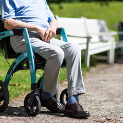 An older adult, sitting in a walker, outside by a bench.