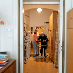 View through a doorway of a younger adult helping an older adult with a walker.