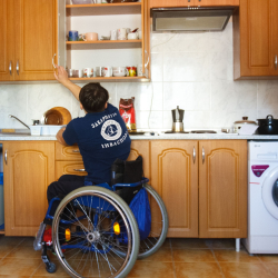 A person in a wheelchair reaching up to get something from a kitchen shelf. 